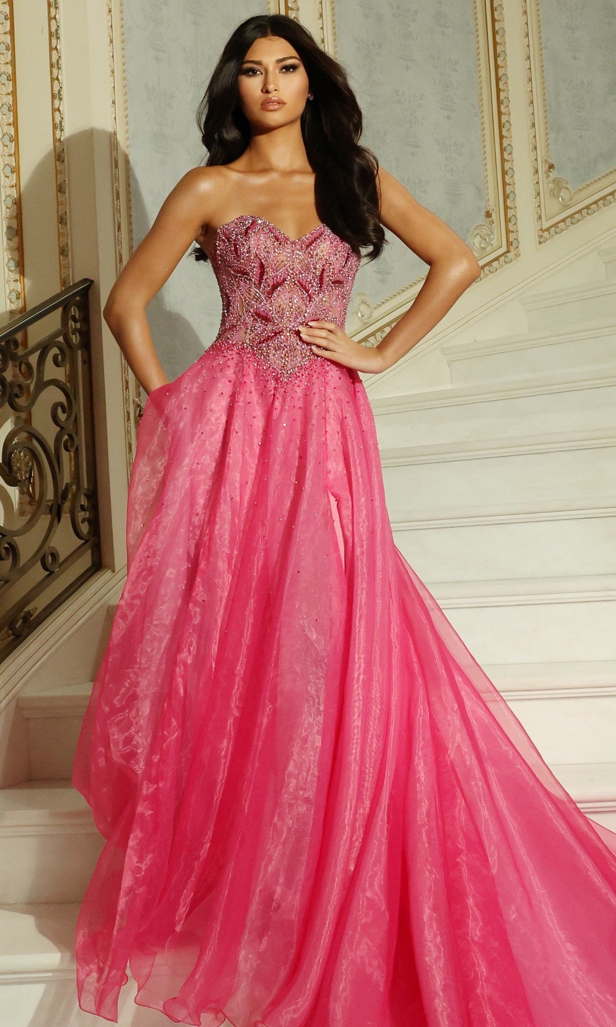 A woman stands on a staircase wearing the Jovani 48906 long prom dress in pink, featuring a strapless beaded corset bodice and a flowing A-line organza skirt.